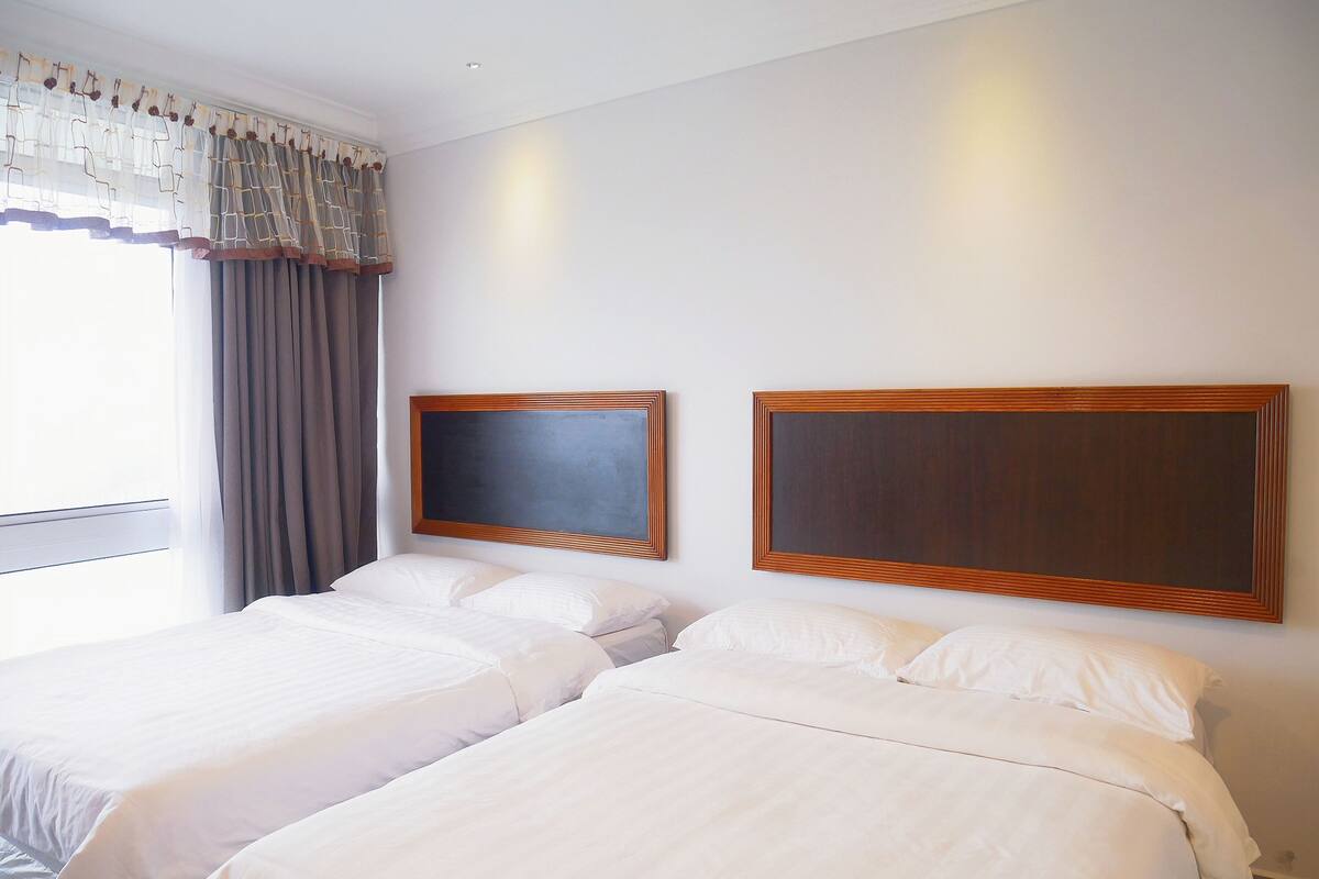 Ein Hotelzimmer mit zwei Betten, einem großen Fenster mit Vorhängen und zwei gerahmten Kunstwerken an der Wand.
