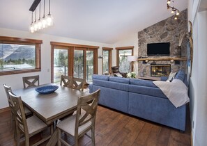 A modern living room with a stone fireplace, a blue sofa, wooden dining table, and a TV mounted on the wall. - Timberfalls 1706 3 Bedroom Condo by RedAwning (Vail)