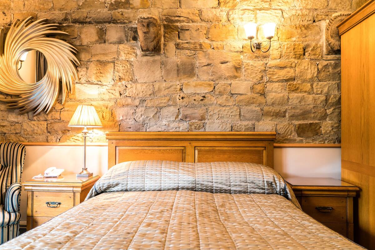 A bedroom with a stone wall, a wooden headboard, a bed with a quilted blanket, a bedside lamp, a mirror, and a nightstand with a telephone.