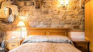 A bedroom with a stone wall, a wooden headboard, a bed with a quilted blanket, a bedside lamp, a mirror, and a nightstand with a telephone.