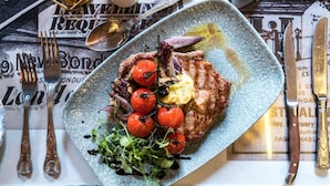 Un plato de comida con filete, tomates cherry y verduras, sobre una mesa con un periódico y cubiertos.