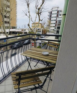 Un balcón con bancos de madera y baranda de metal, con vista a una calle con coches estacionados y edificios.