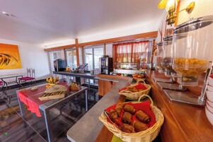 Zona de desayuno tipo buffet con diferentes opciones de alimentos, una máquina de café y una sala de estar con mesa y sillas.