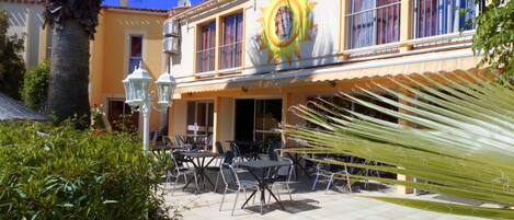 Un café al aire libre con mesas y sillas, una palmera y un edificio con un mural de sol.