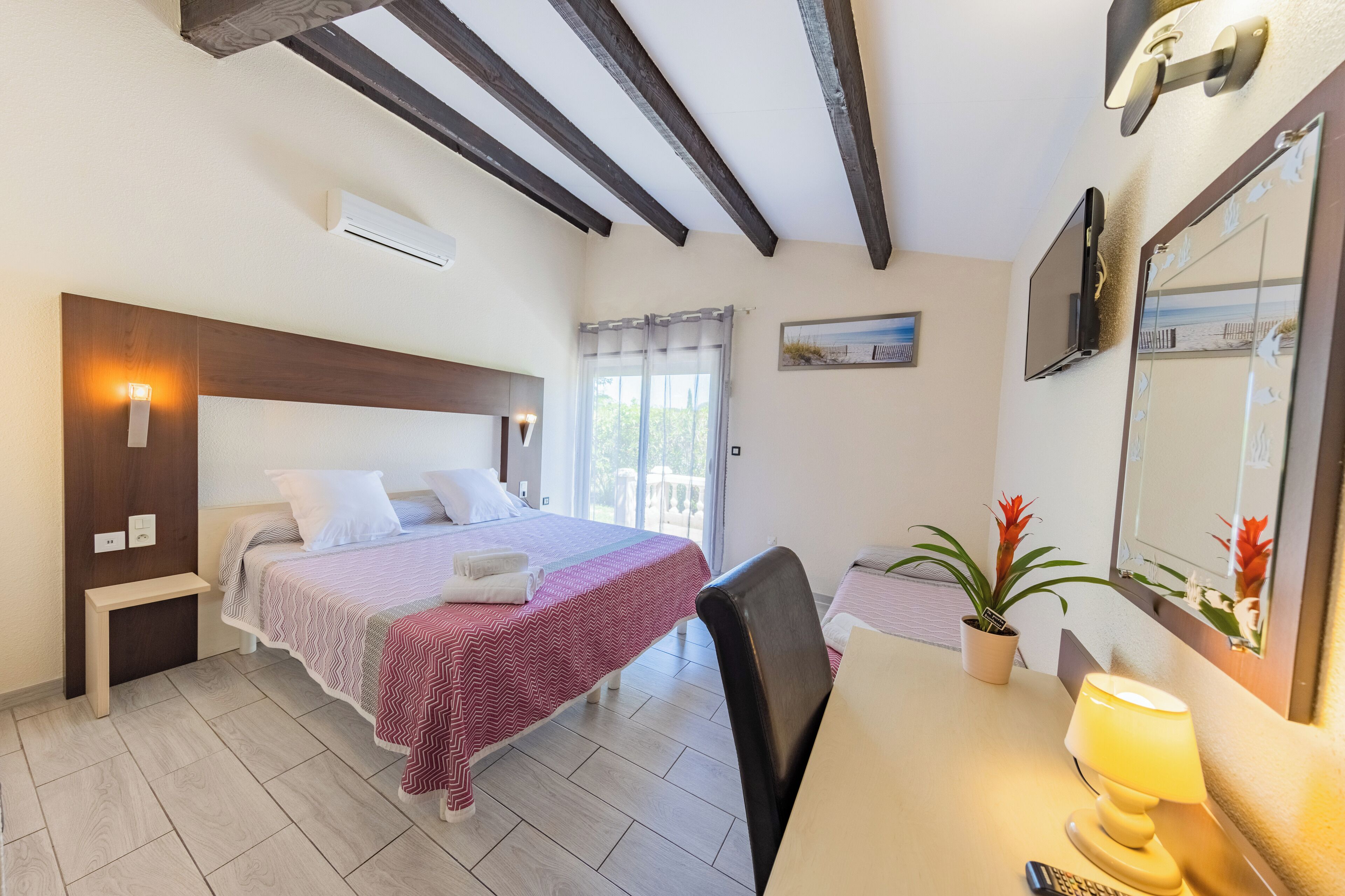 A bedroom with a bed, bedside table, lamp, potted plant, and a window with curtains.