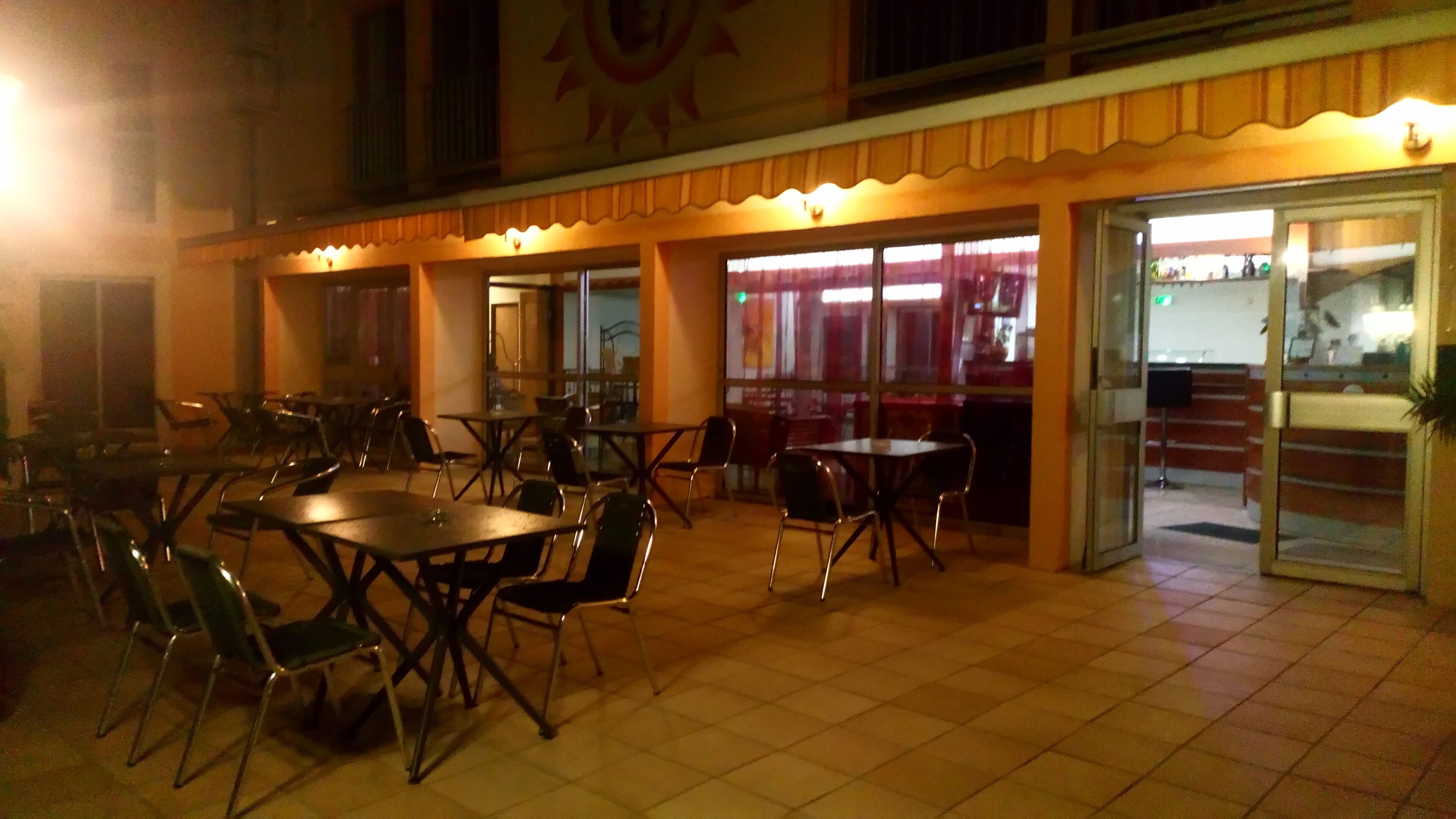 An outdoor dining area with tables and chairs, a tiled floor, and a building entrance with glass doors.