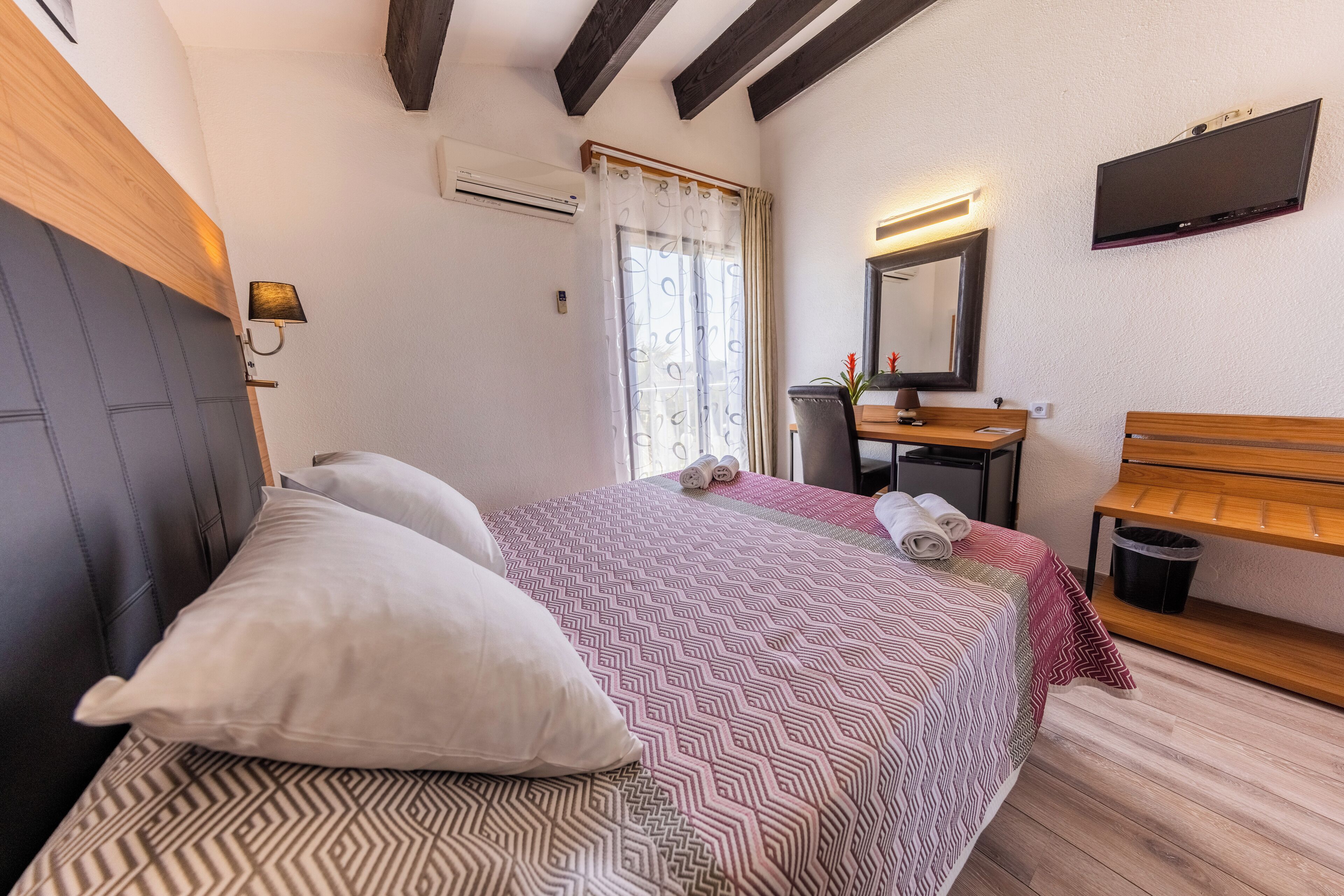 Un dormitorio con cama, almohadas, un banco de madera, un espejo, un televisor y una ventana con cortinas.