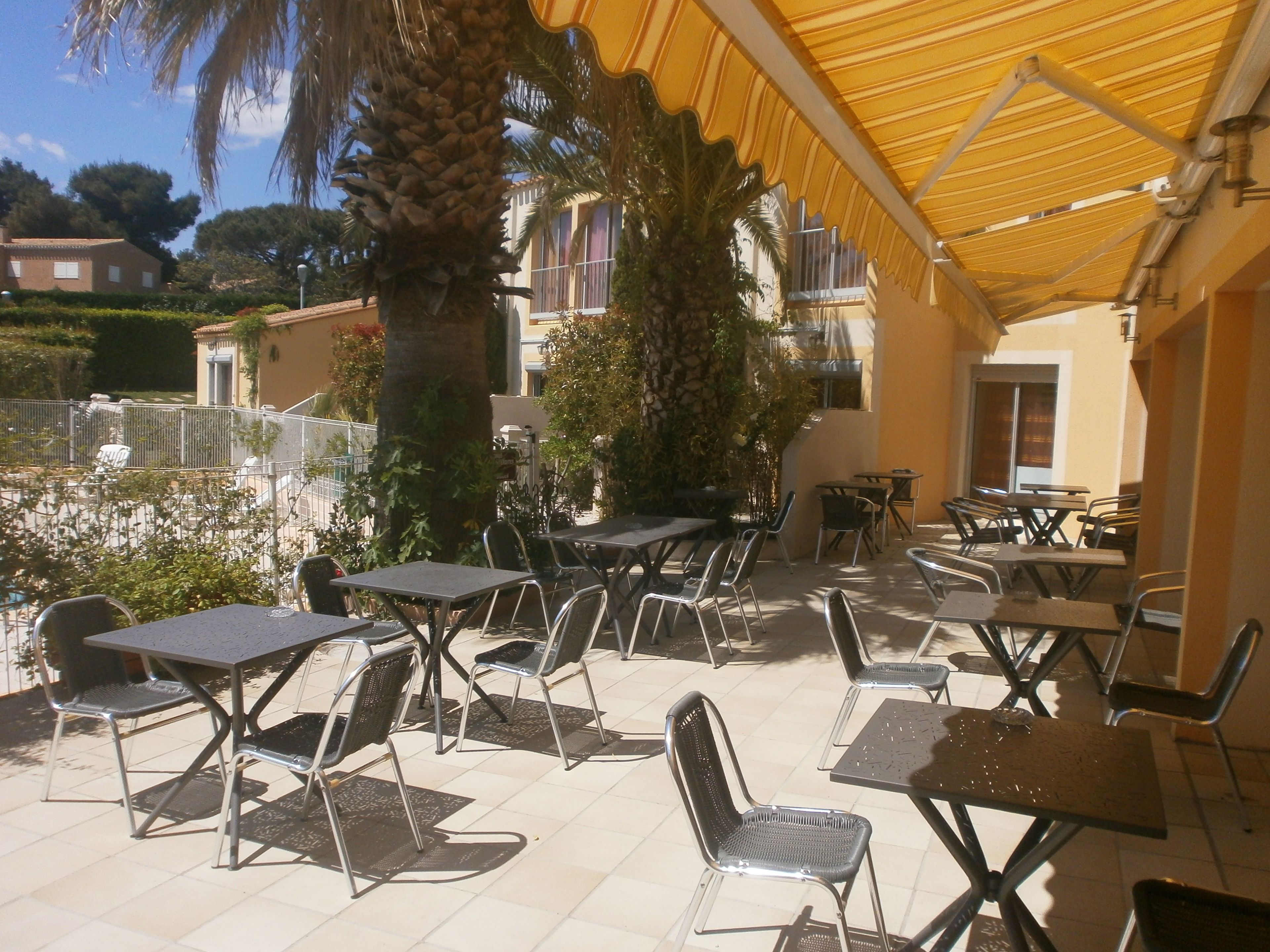 An outdoor patio area with tables and chairs, surrounded by palm trees and a yellow canopy.