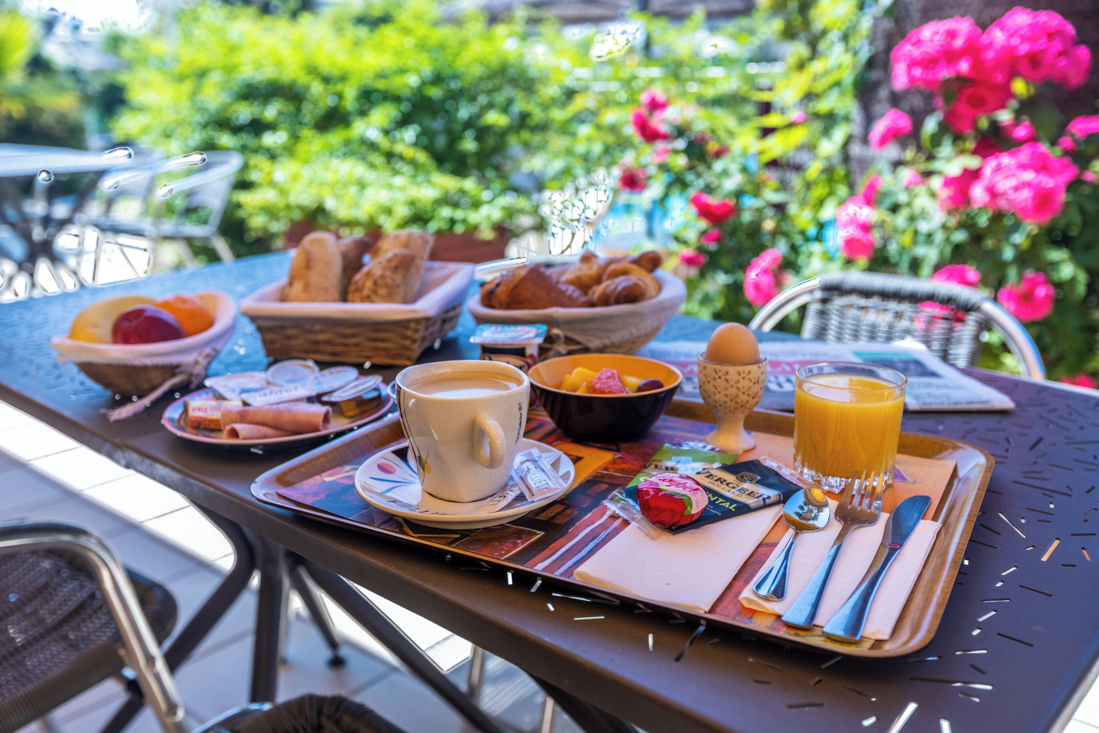 Un plateau de petit-déjeuner avec des croissants, des fruits, du café et du jus, placé sur une table à l’extérieur, avec des chaises et de la verdure en arrière-plan.