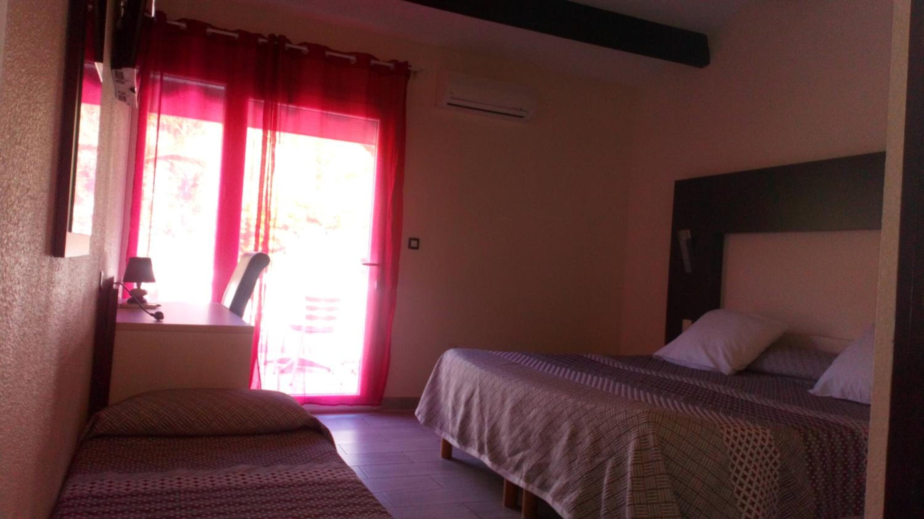 Ein Zimmer mit zwei Betten, einem Schreibtisch mit Lampe und einem Fenster mit roten Vorhängen.