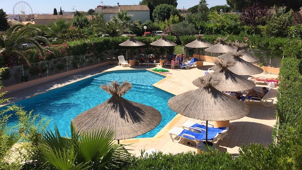 Una piscina con sillas de descanso y sombrillas, rodeada de vegetación y una noria al fondo.