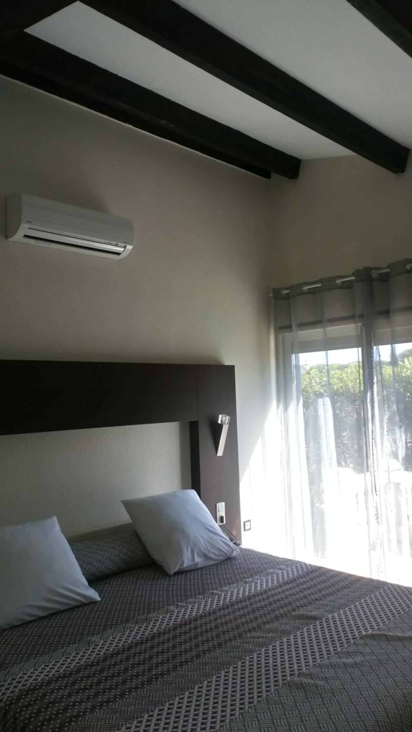 Une chambre d’hôtel avec un lit, une tête de lit, un appareil de climatisation, une fenêtre avec des rideaux et une vue sur la verdure.