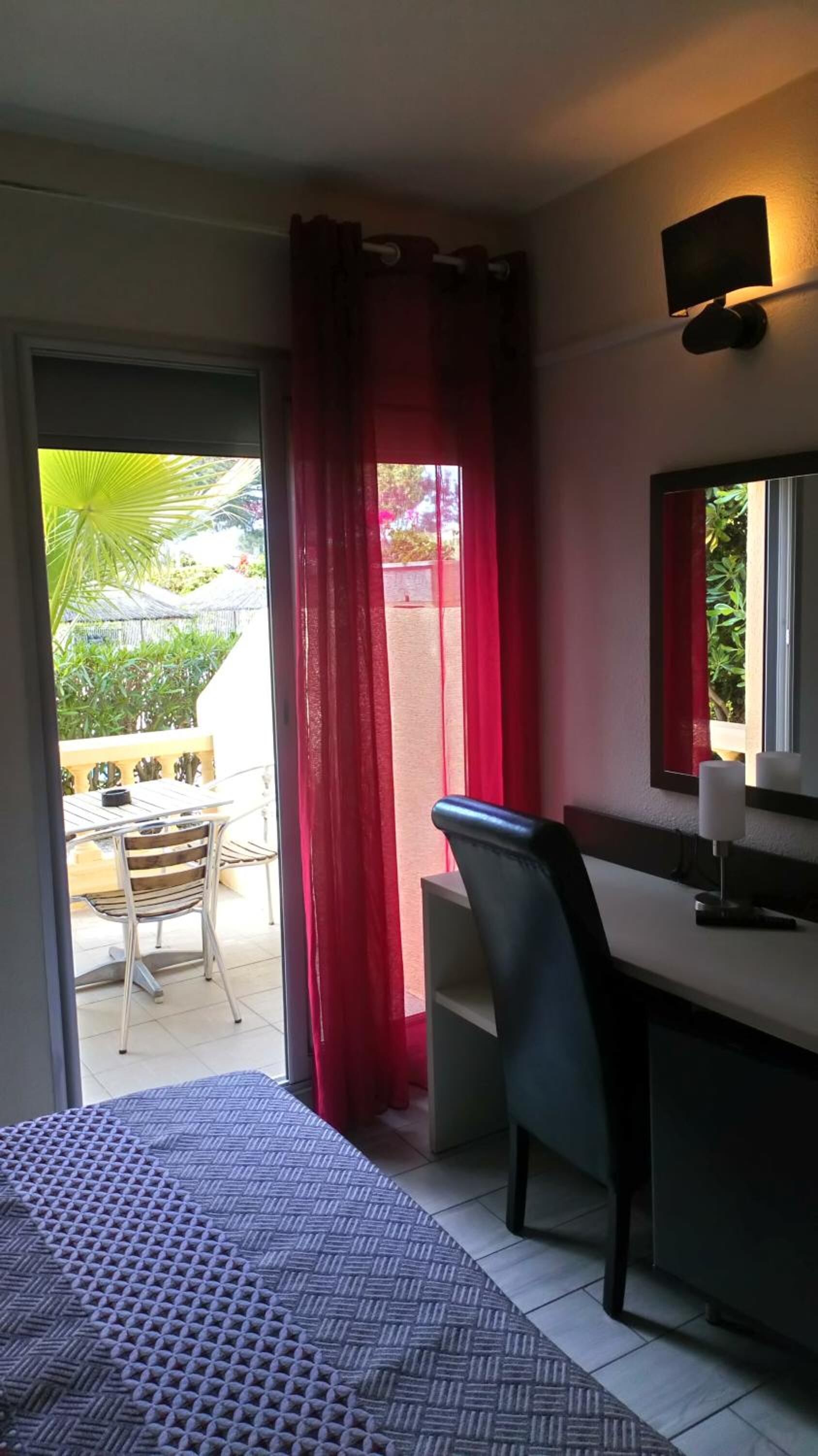 Une pièce comprenant un lit, un bureau et une chaise. Il y a une fenêtre avec des rideaux rouges, et la vue donne sur une terrasse avec une table et des chaises.