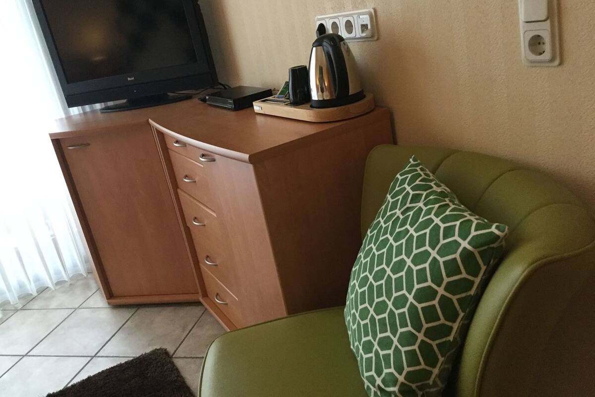Une pièce avec un téléviseur posé sur un meuble en bois, un canapé vert agrémenté d’un coussin à motifs et une machine à café sur le meuble.