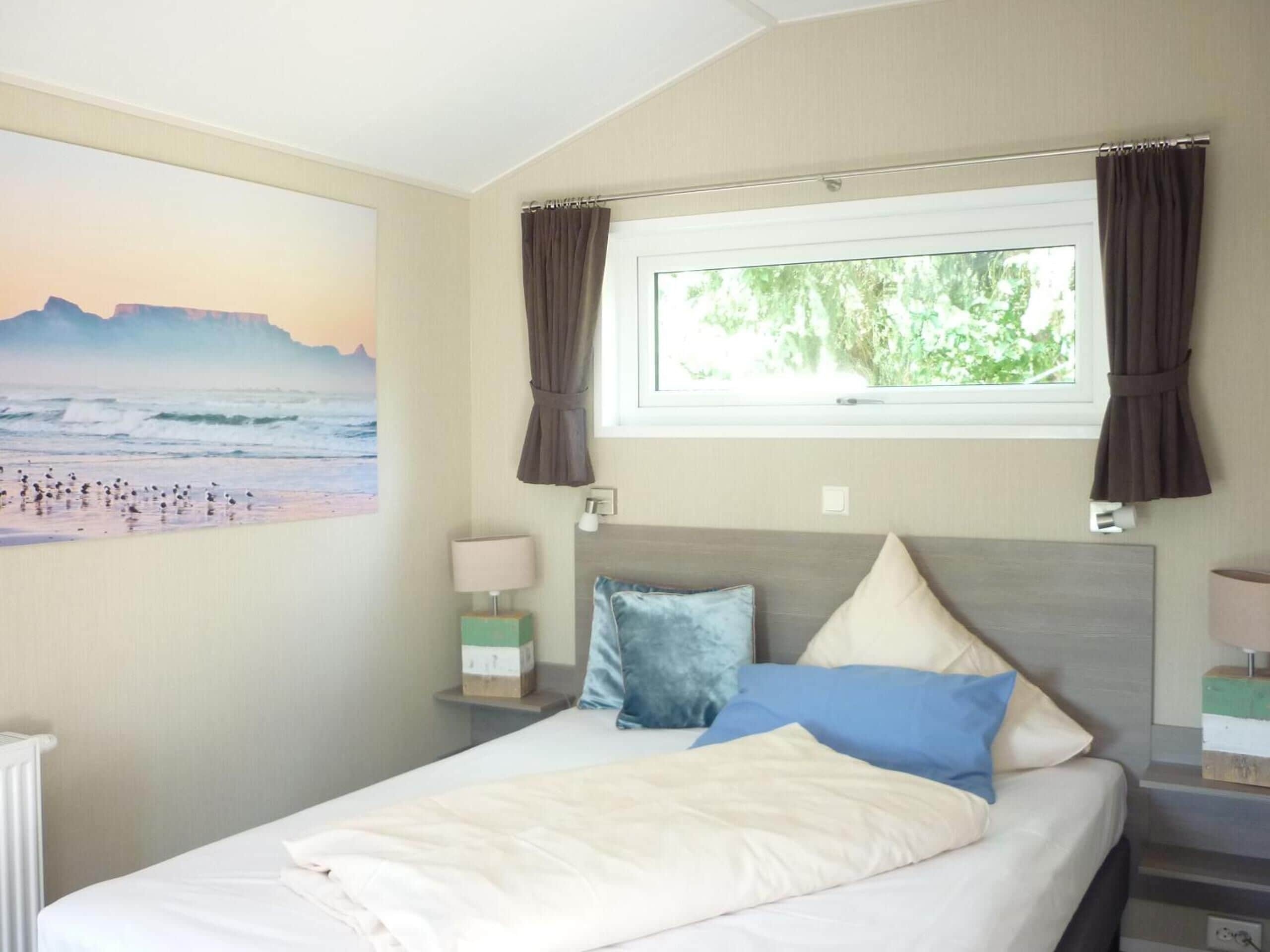 A bedroom with a bed, a window with curtains, a wall-mounted lamp, and a painting of a beach scene.