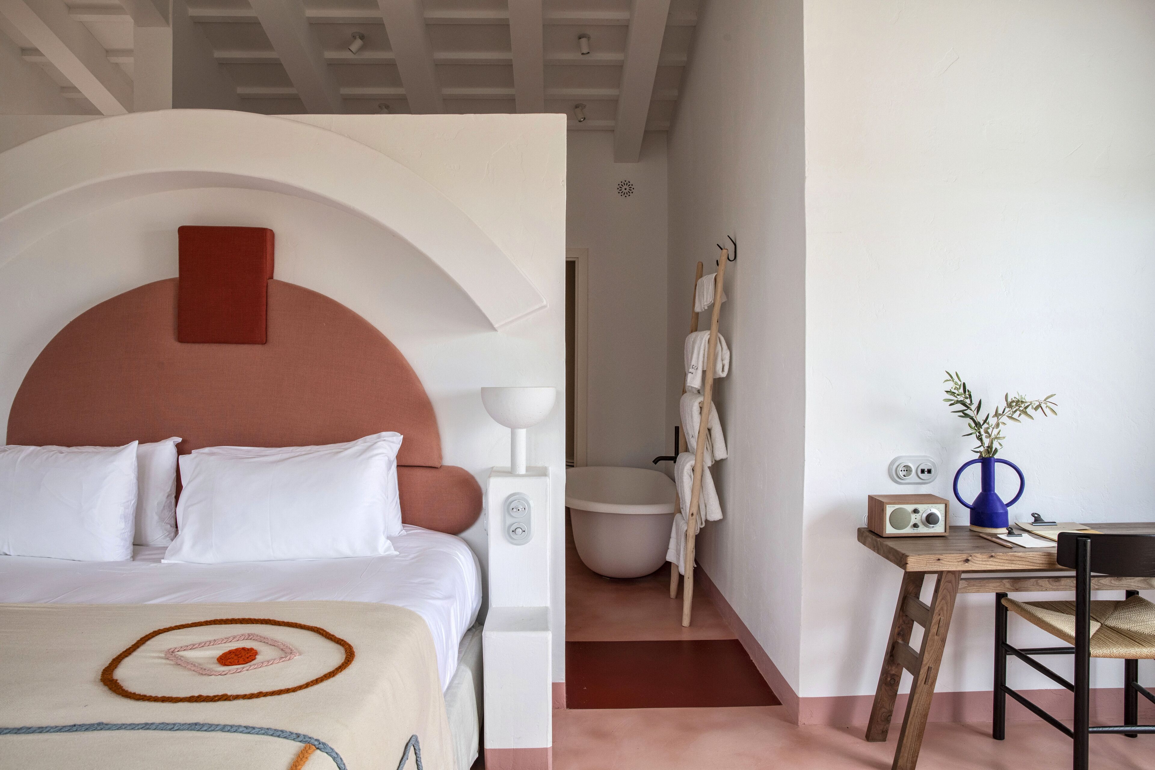 A hotel room with a bed, a desk, a radio, and a vase with plants.