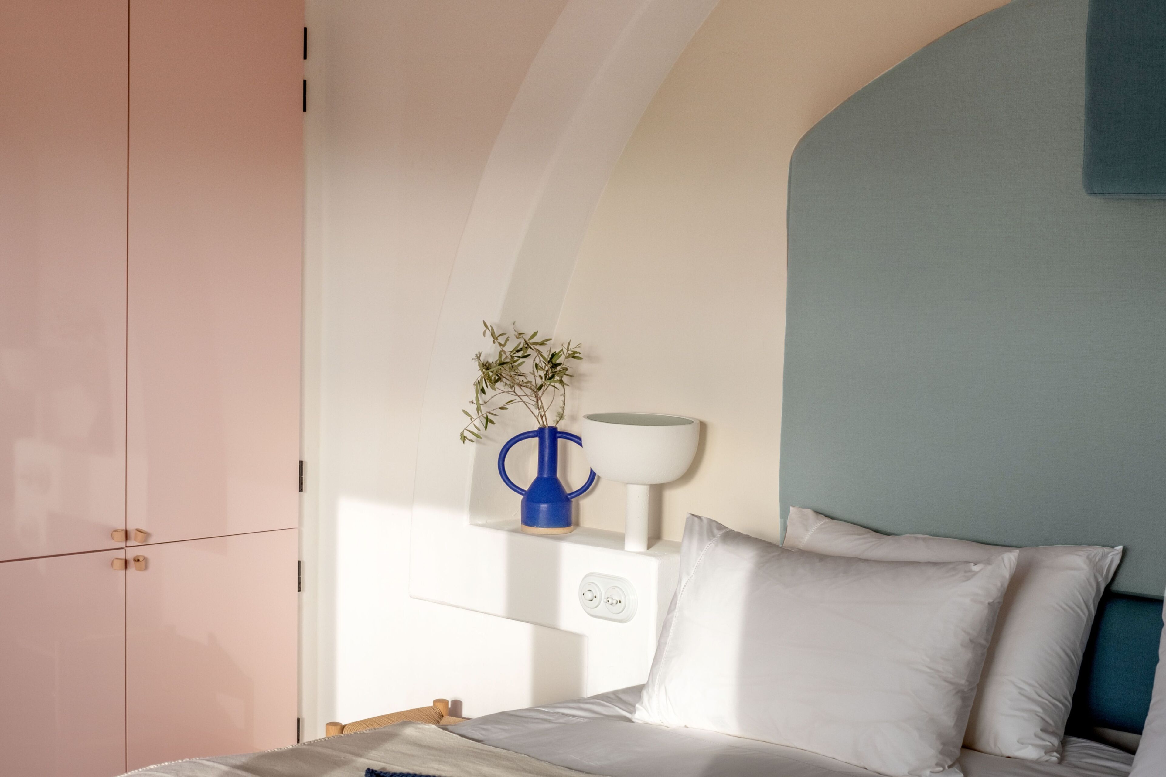 A bedroom with a bed, a nightstand, a vase with a plant, and a wall-mounted lamp.