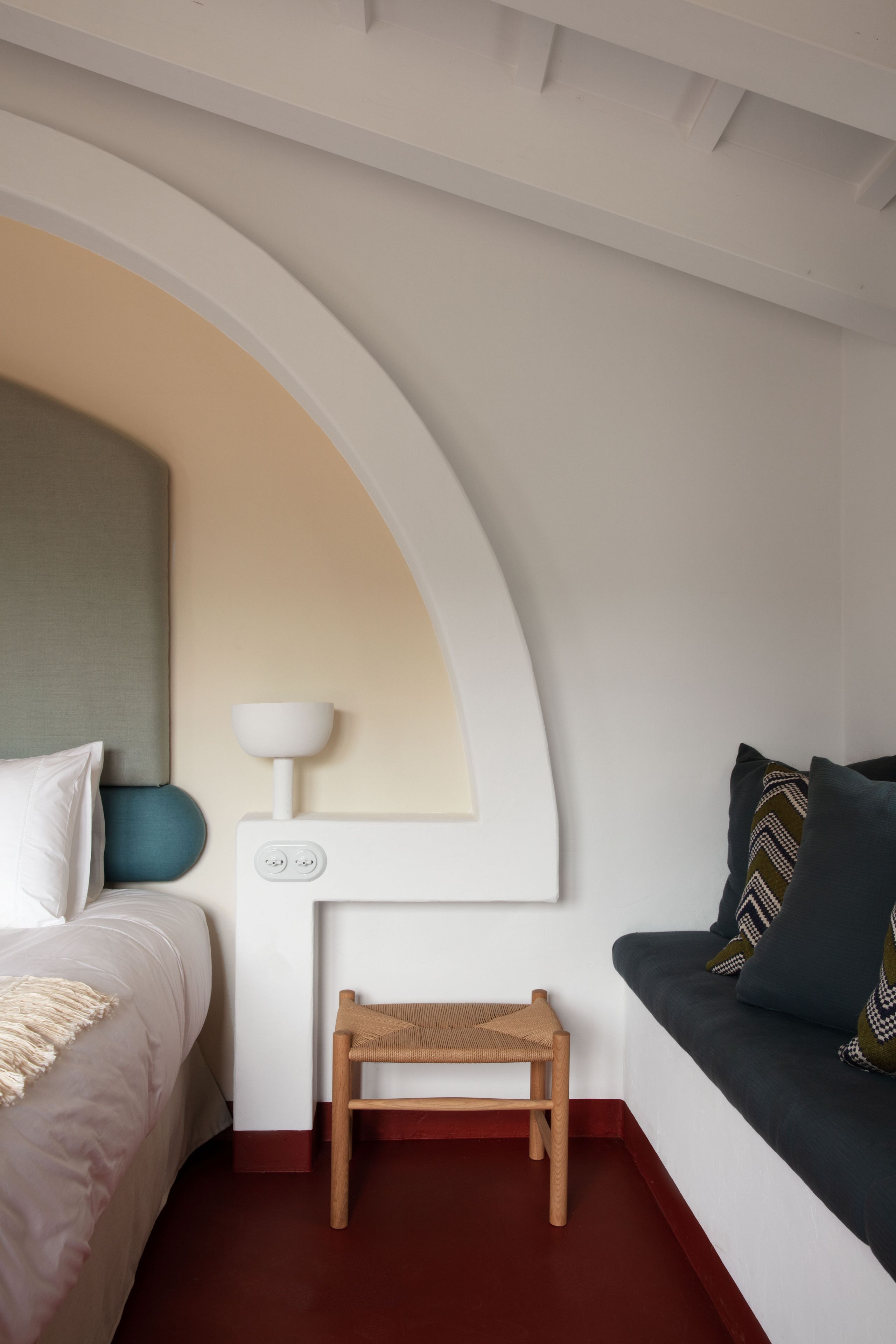 A modern bedroom with a curved ceiling, a bed with white bedding, a small wooden stool, a white nightstand with a lamp, and a grey sofa with patterned cushions.