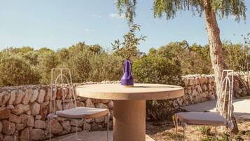 Un espacio al aire libre con una mesa redonda y dos sillas, rodeado por un muro de piedra y árboles.