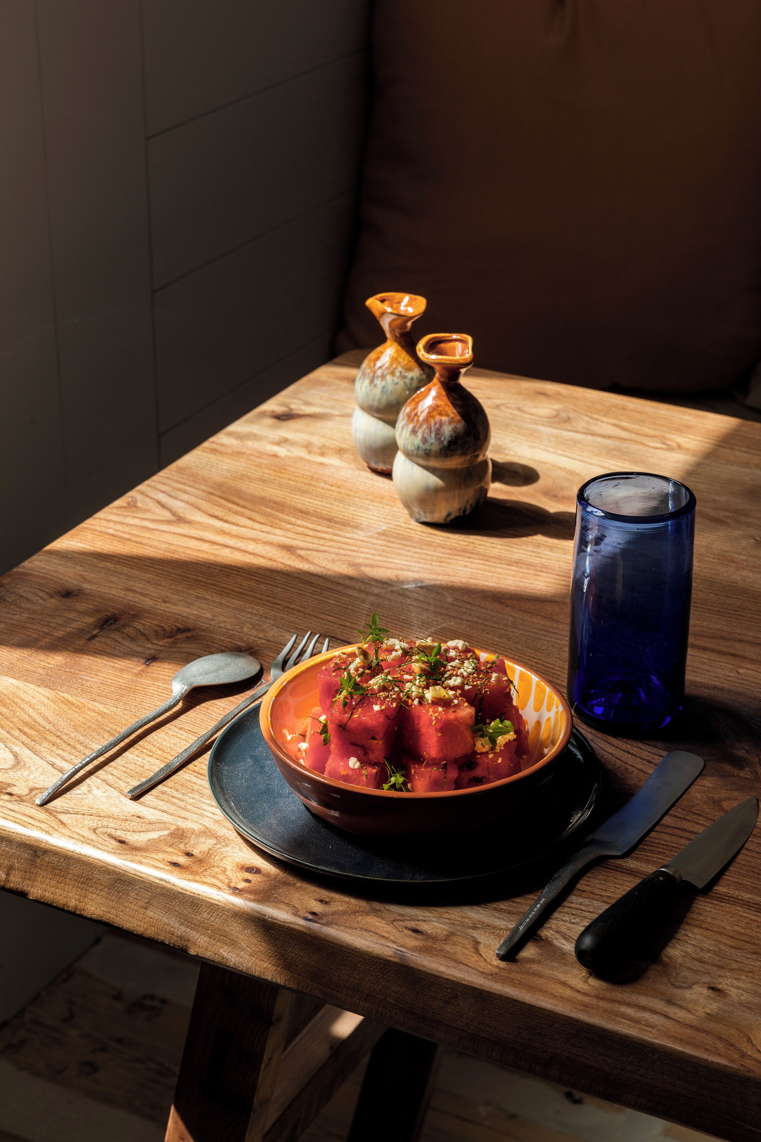 A wooden table with a bowl of food, a blue glass, and cutlery.