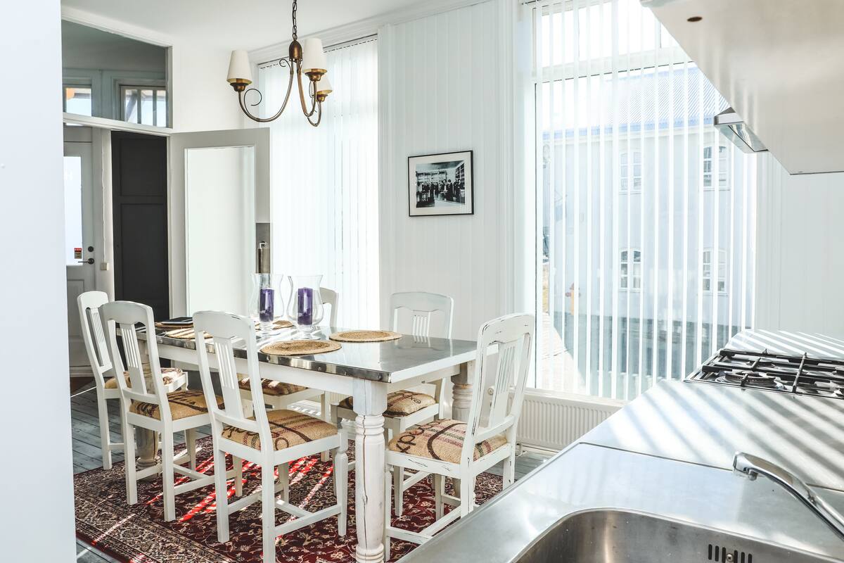 A dining area with a white table and chairs, a chandelier, and a large window with blinds.