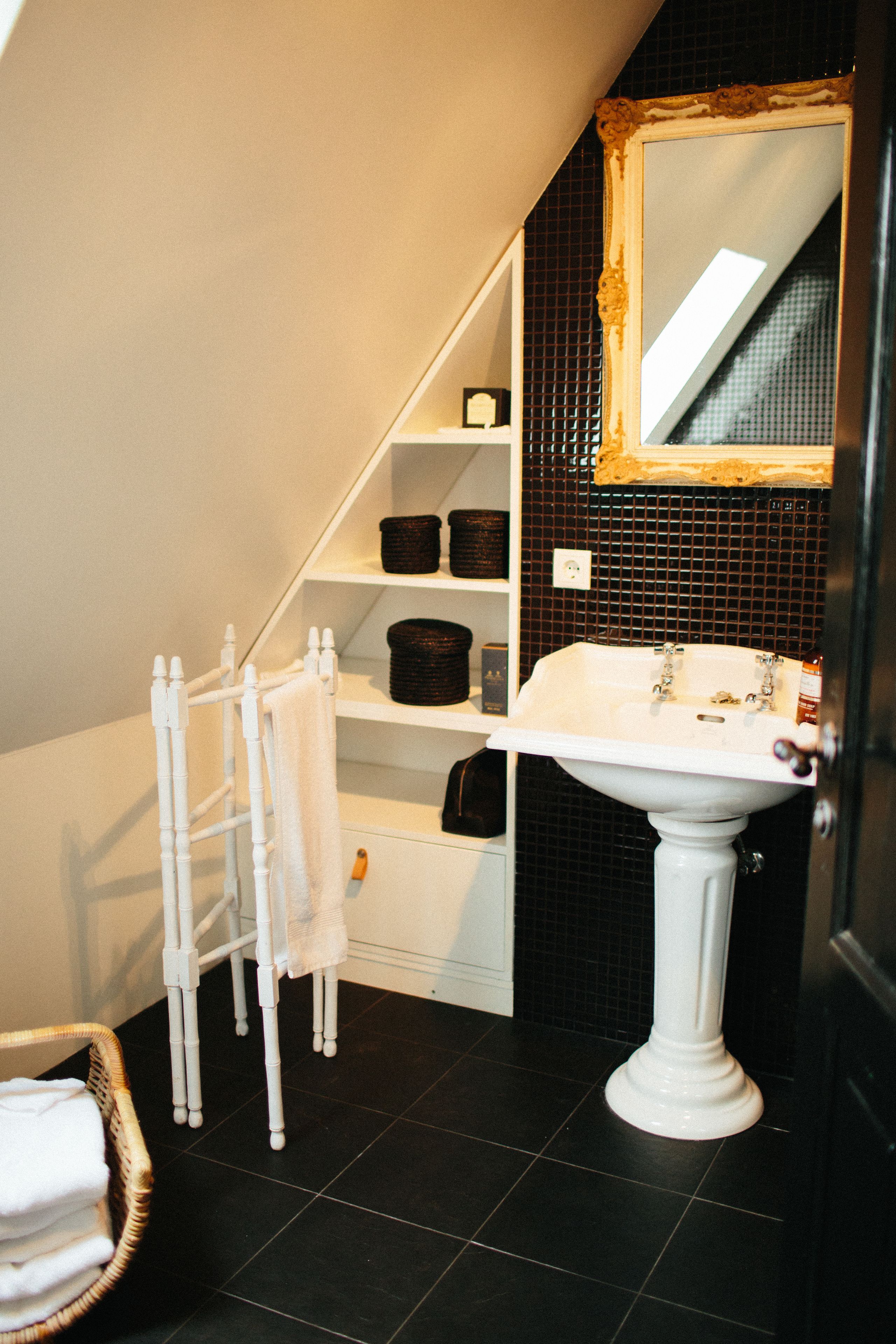 A bathroom with a white pedestal sink, a mirror with a gold frame, a towel rack, and shelves with baskets.