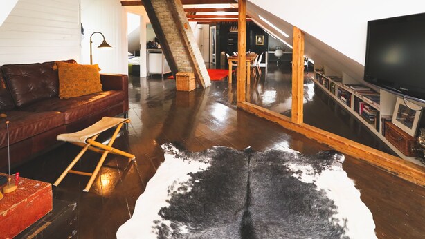 A spacious loft with a high ceiling, exposed wooden beams, a leather sofa, a large flat-screen TV, and a cowhide rug. - Hoepfner  Historical House (Akureyri)