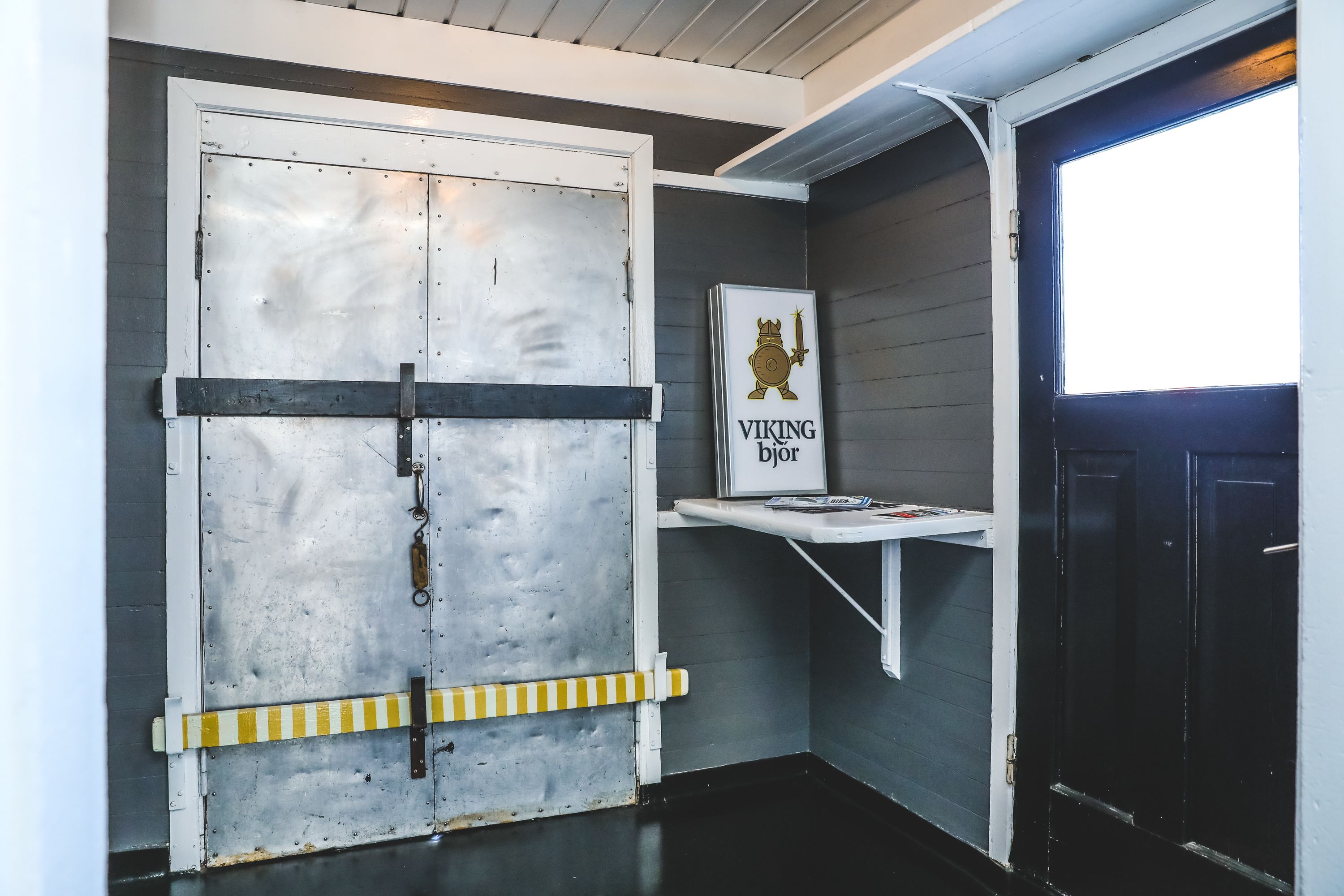 A room with a large metal door, a small shelf with a sign, and a window.