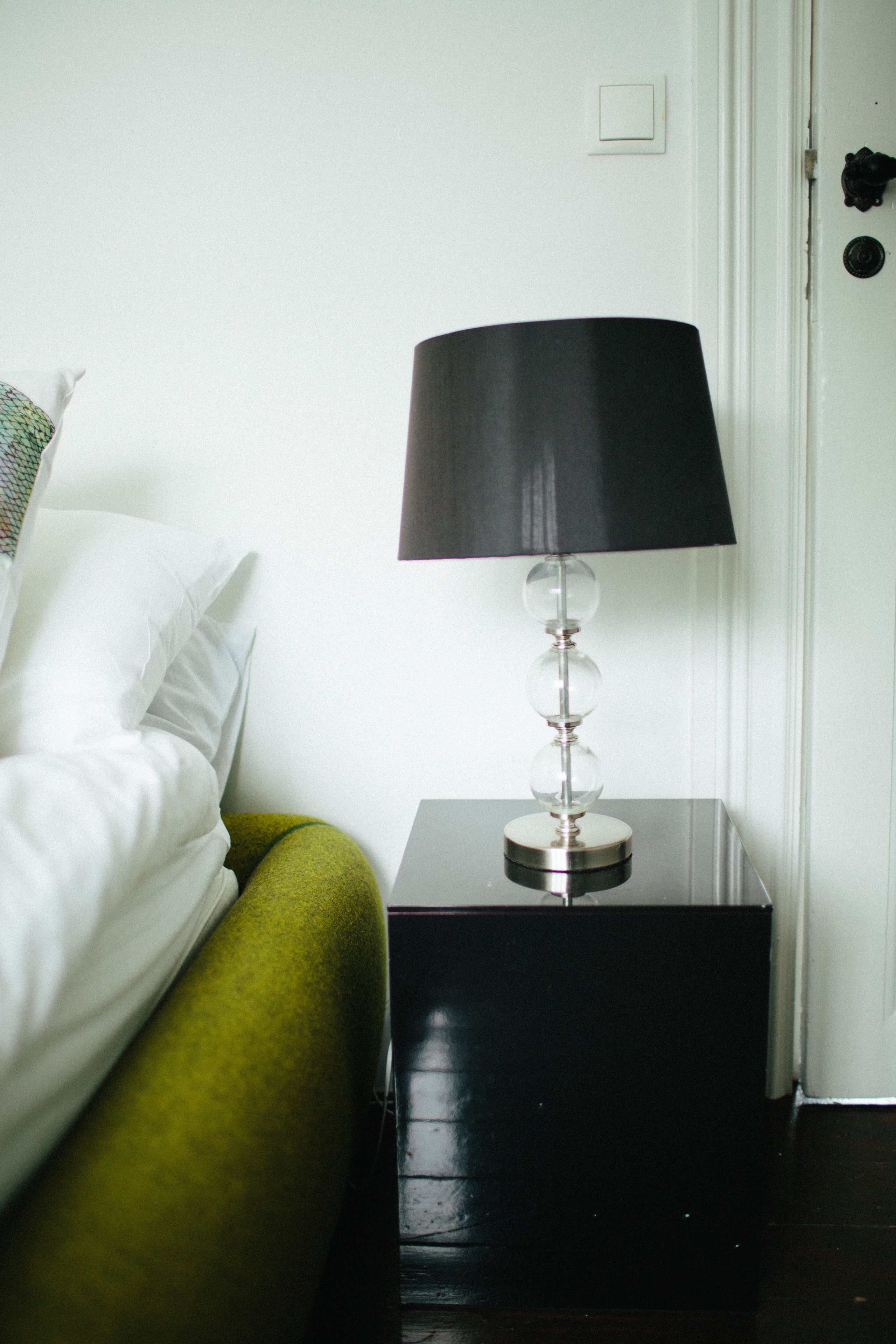 A neatly made bed with white linens and a green accent, a black lamp on a small table beside it, and a white door in the background.