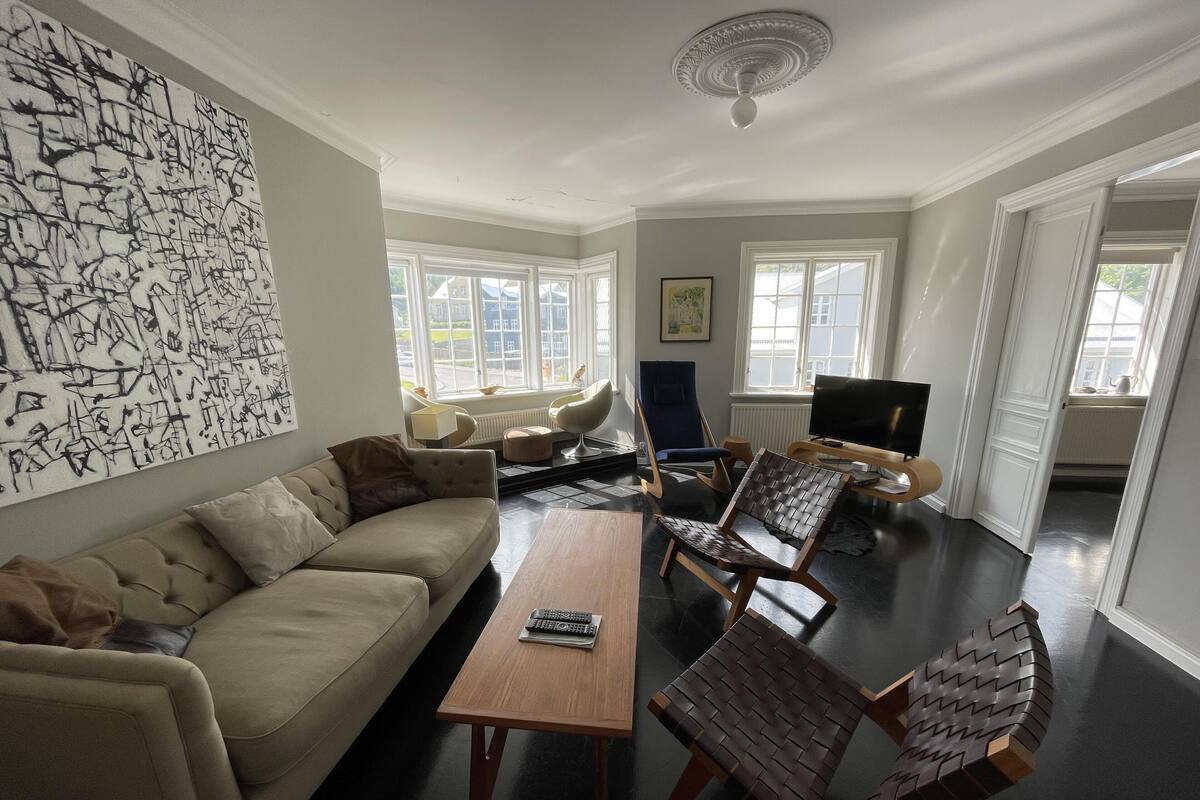 A living room with a sofa, armchairs, a coffee table, and a TV. There are windows with white frames and a ceiling fan.
