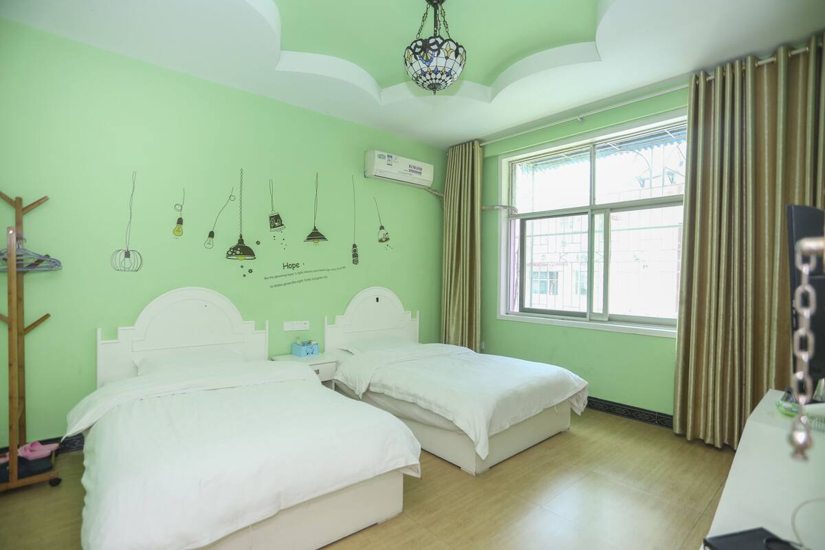 A room with two beds, a chandelier, a window with curtains, and a wall-mounted air conditioner.