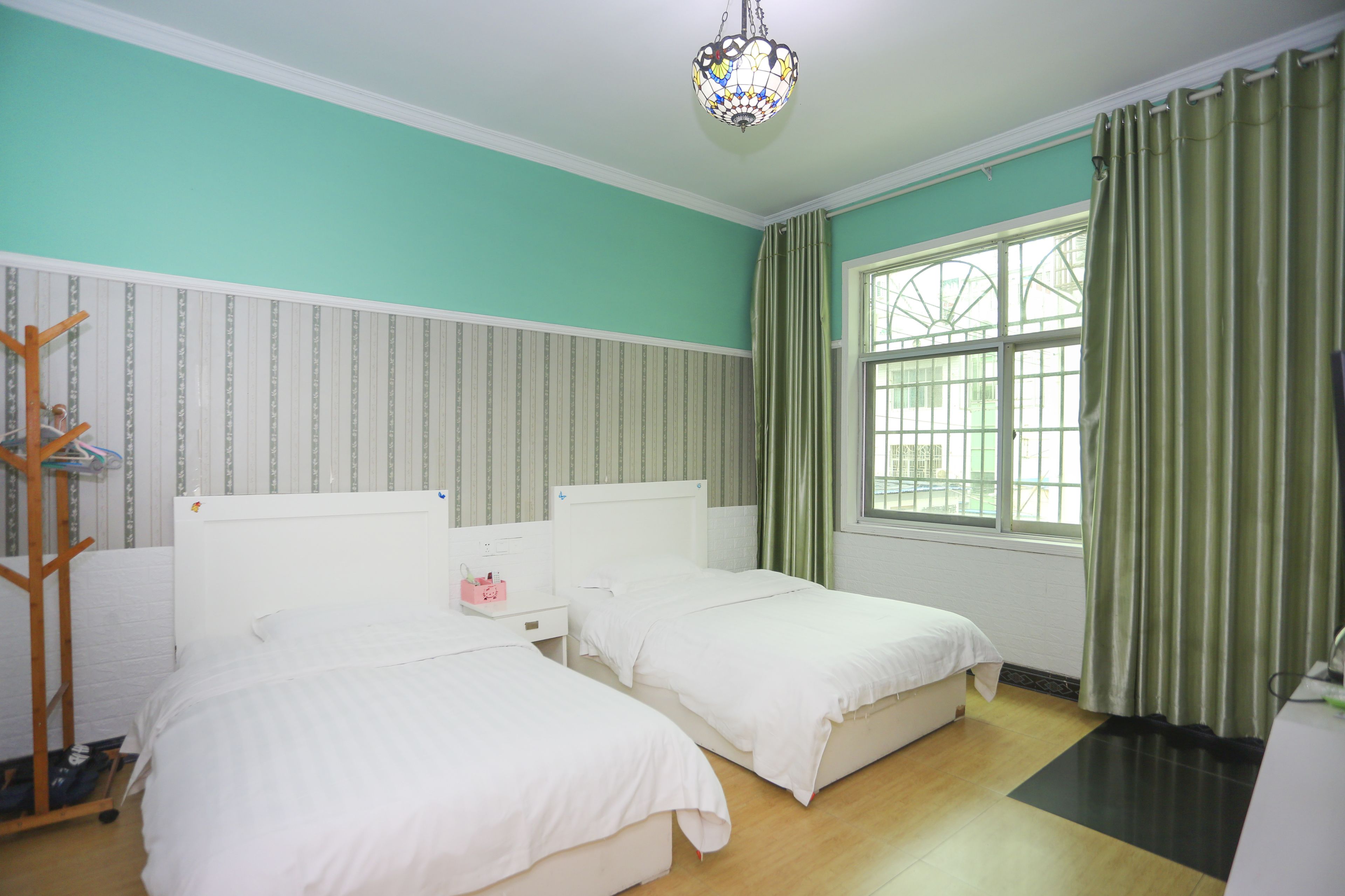 A hotel room with two single beds, a wooden coat rack, a bedside table with a pink object, and a large window with green curtains.