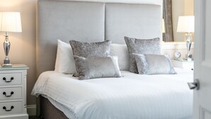 Cama bem arrumada com cabeceira cinza, roupa de cama branca e almofadas cinza estampadas. Um criado-mudo branco com abajur e gaveta.