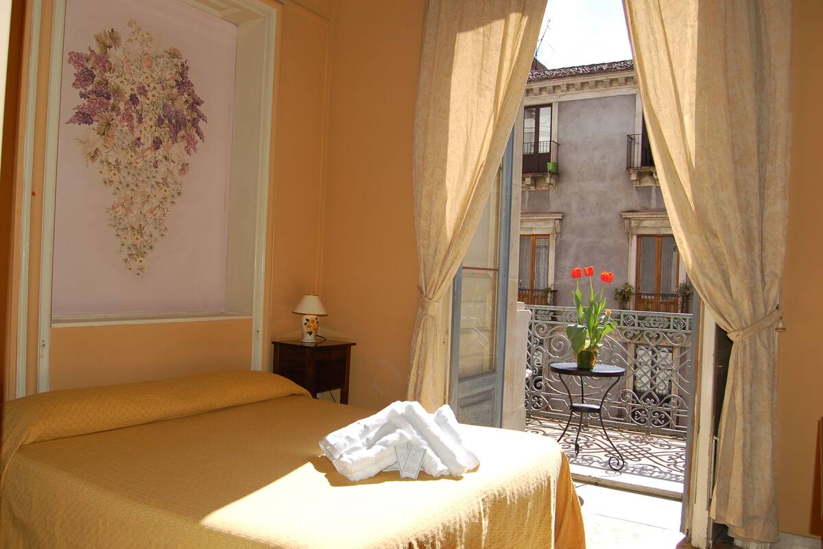 A hotel room with a bed, a nightstand, a window with curtains, and a balcony with a potted plant and red flowers.