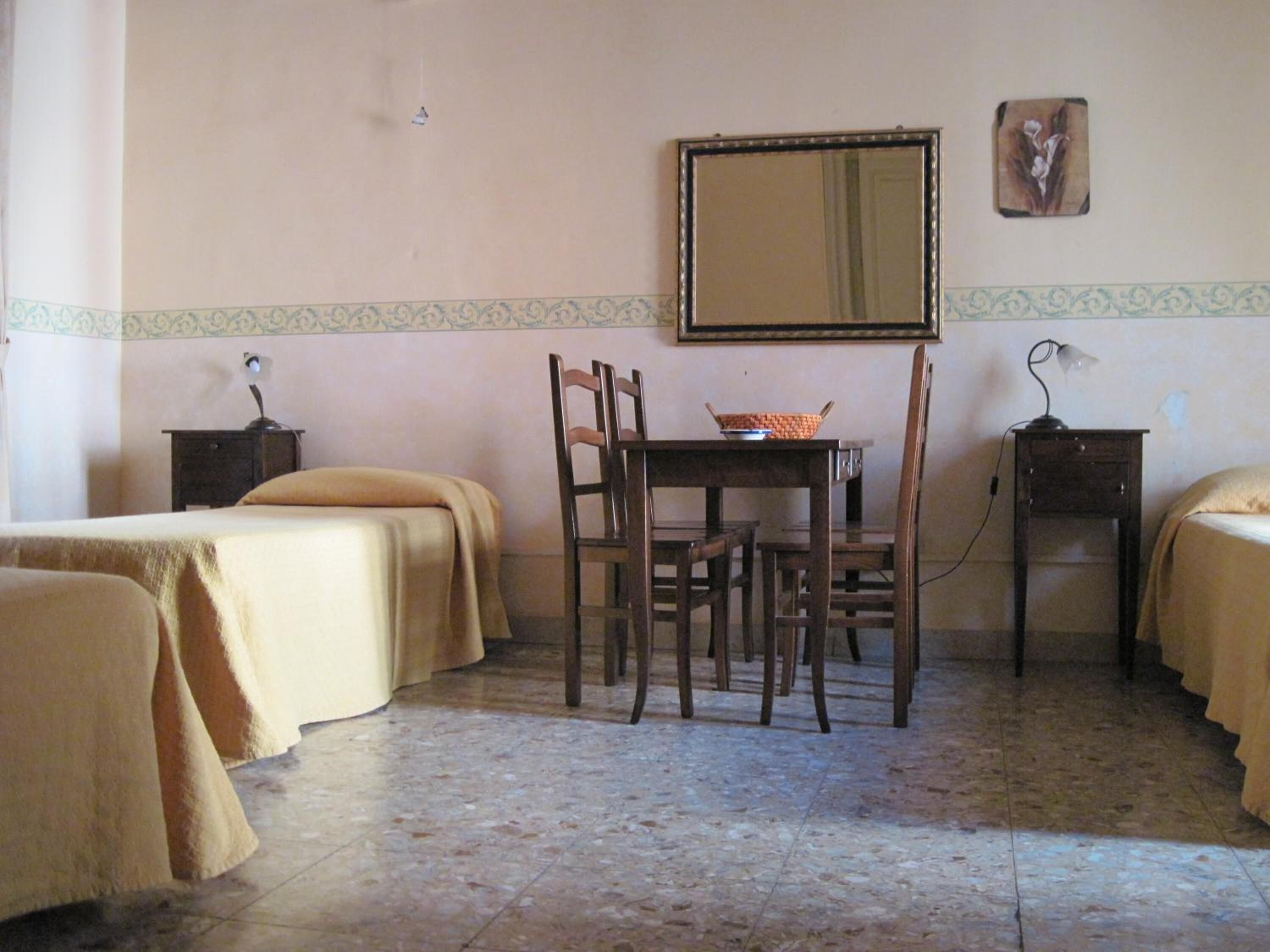 A room with two single beds, a wooden table with chairs, a small table with a lamp, and a wall-mounted mirror.