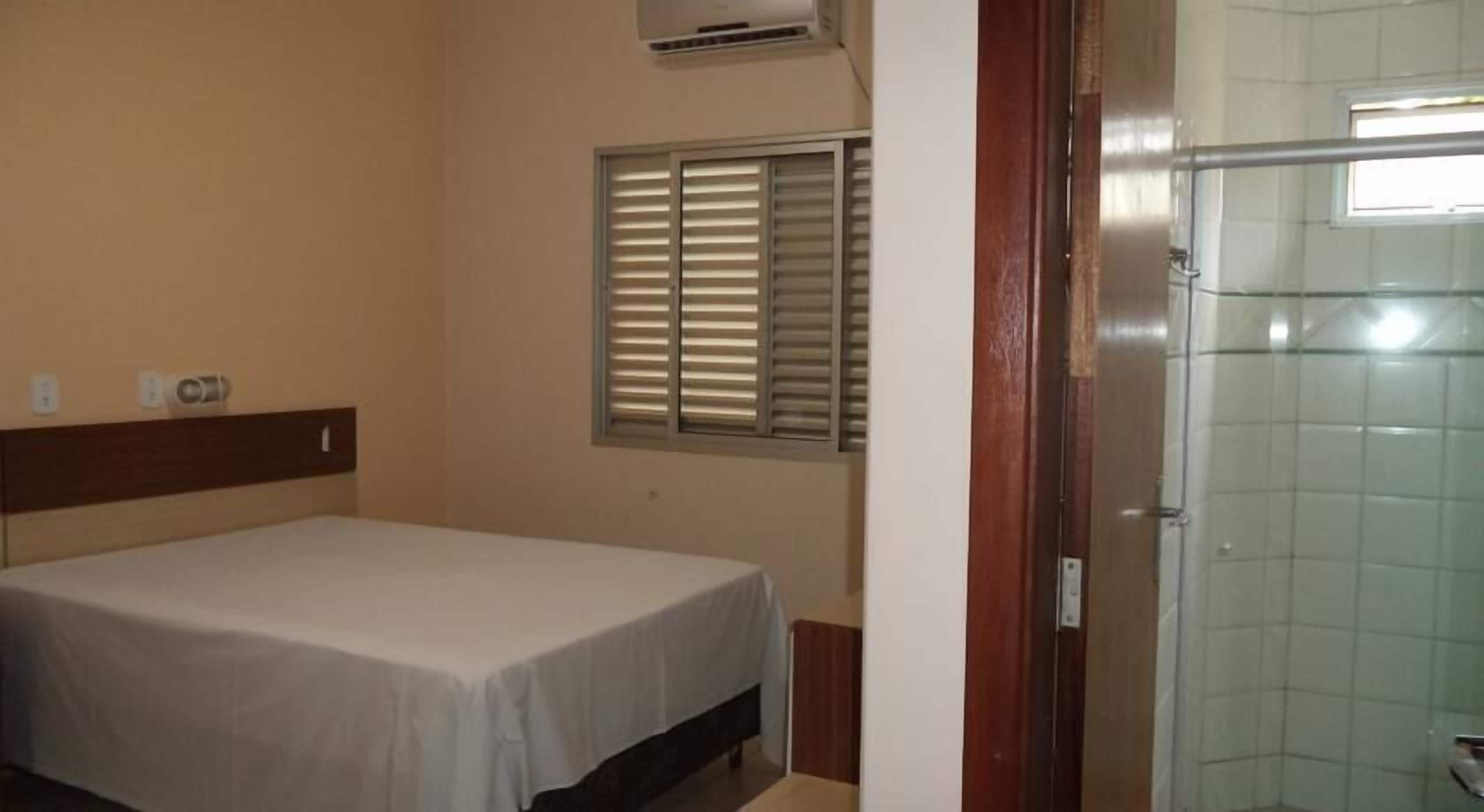 Quarto com cama, janela com persianas, área de chuveiro e porta.