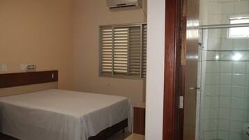 Quarto com cama, janela com persianas, área de chuveiro e porta.