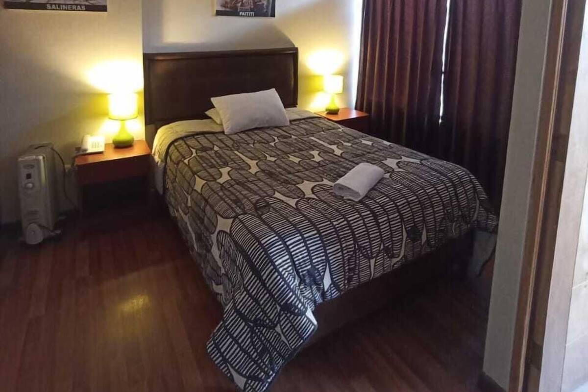 A hotel room with a large bed, two bedside lamps, a nightstand with a lamp, a wall-mounted heater, and a window with curtains.