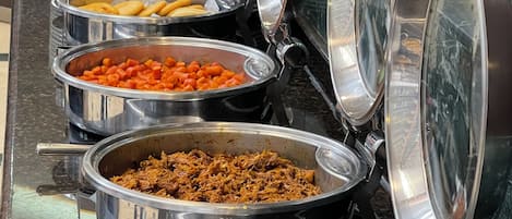 A buffet with various food items in metal serving dishes, including a dish with what appears to be shredded meat, a dish with cubed carrots, and a dish with chocolate chips.
