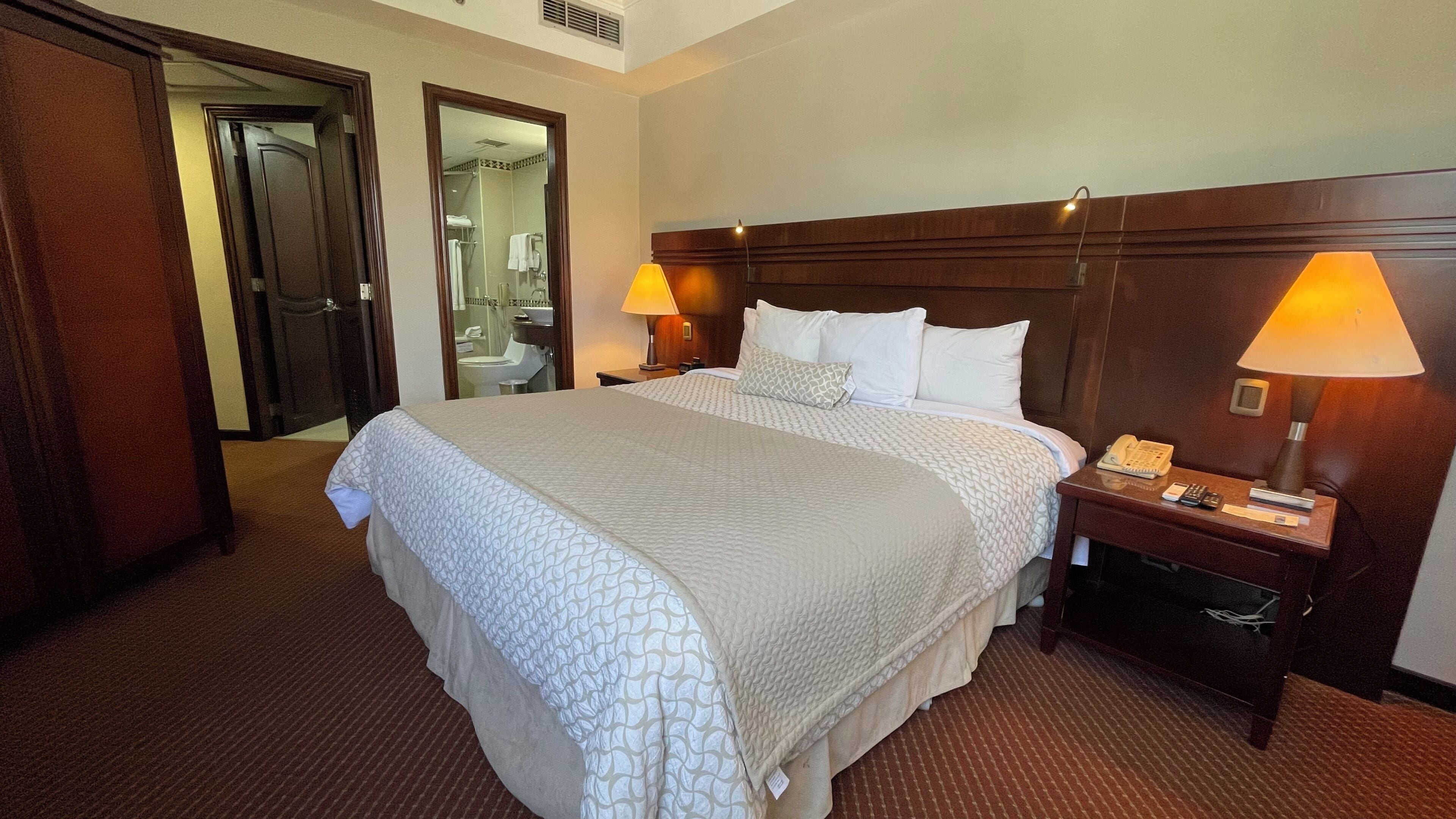 A hotel room with a large bed, two bedside lamps, a nightstand with a phone, and a mirror reflecting a bathroom.