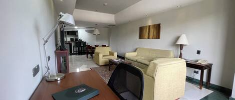 A modern living room with a sofa, armchairs, a desk with a laptop, and a dining area in the background.