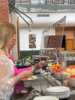 Zona de bufé con diferentes opciones de comida, una persona sirviendo y una pared de ladrillo al fondo.