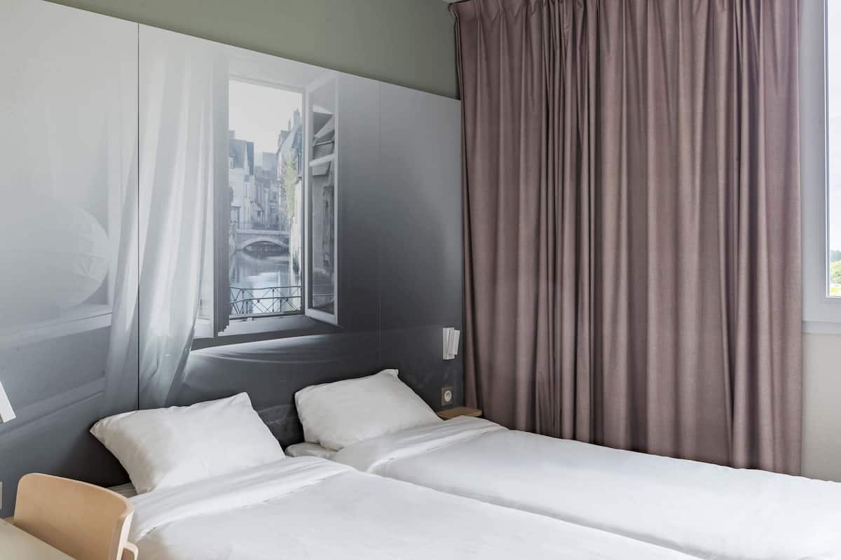 A hotel room with a bed, a chair, a window with curtains, and a view of a cityscape.