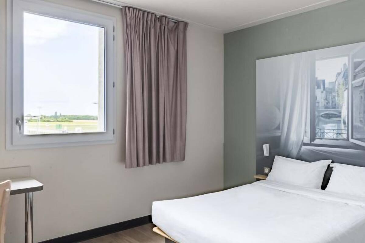 A hotel room with a bed, a window with curtains, a painting on the wall, and a small table.
