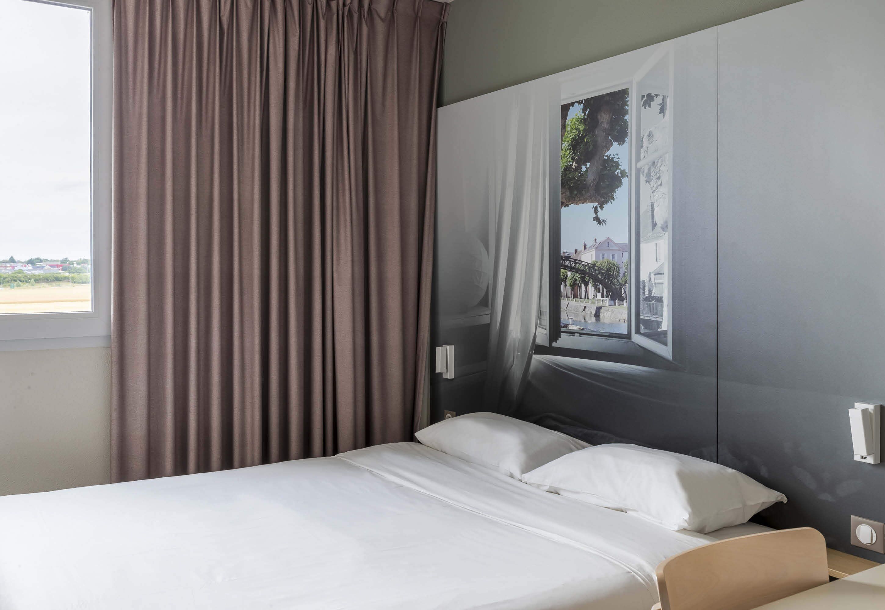 Une chambre d’hôtel avec un lit, une fenêtre avec des rideaux et une œuvre d’art accrochée au mur.
