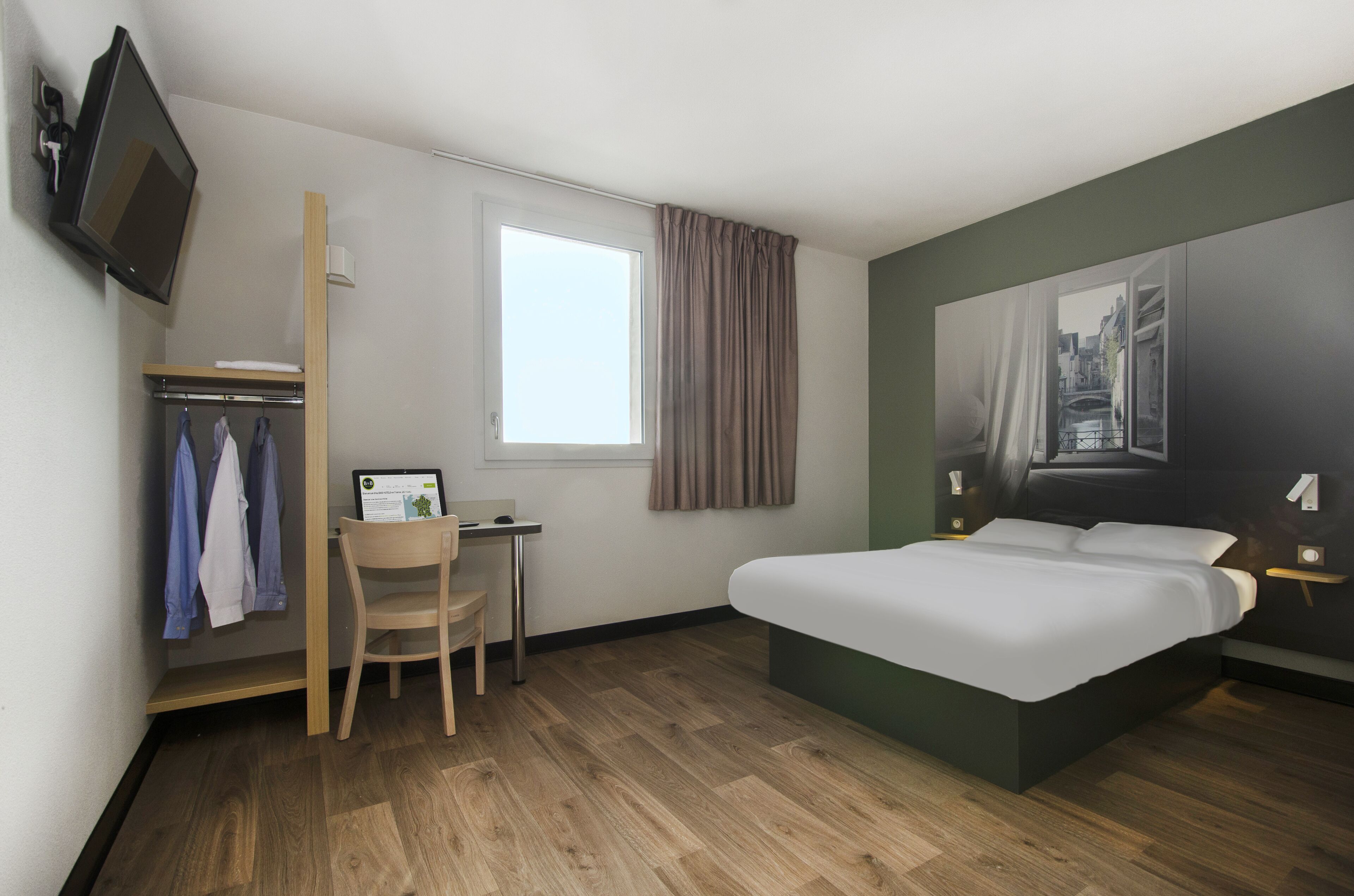 A hotel room with a bed, a desk with a computer, a chair, a wardrobe, a window with curtains, and a TV mounted on the wall.
