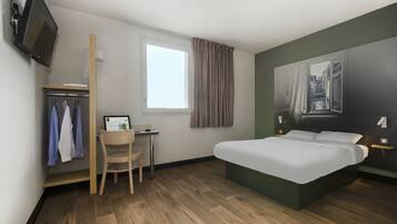 A hotel room with a bed, a desk with a computer, a chair, a wardrobe, a window with curtains, and a TV mounted on the wall.