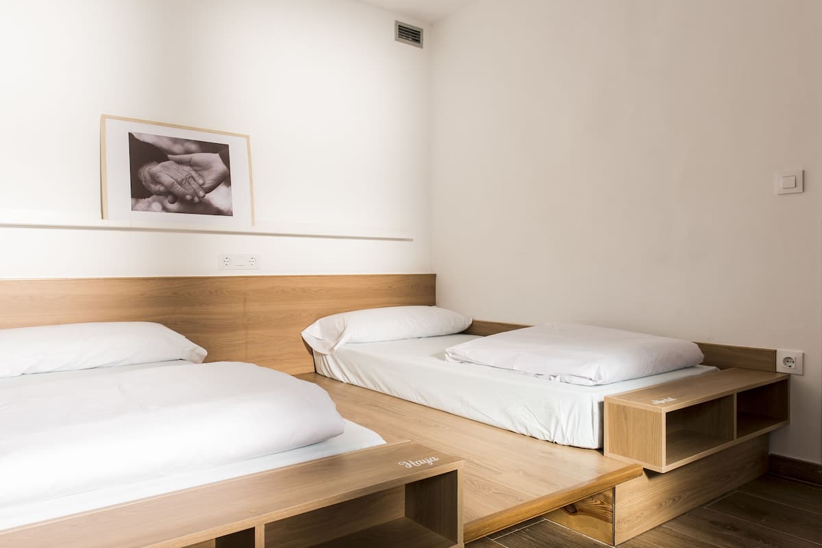 A room with two wooden beds, white bedding, a framed photograph on the wall, and a small wooden shelf.