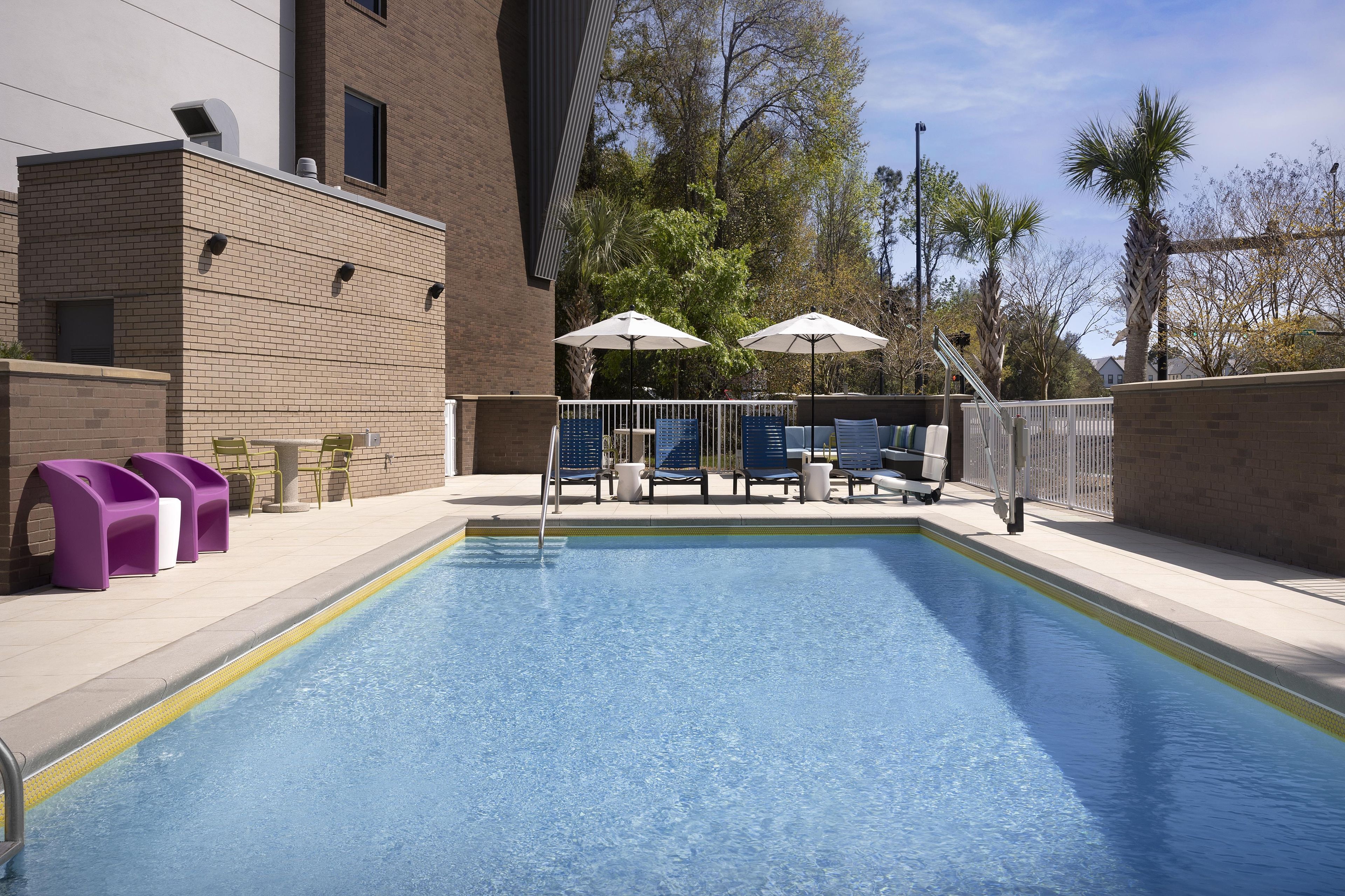 Une piscine entourée d’un bâtiment en briques et d’arbres, avec des chaises longues et des parasols à proximité.