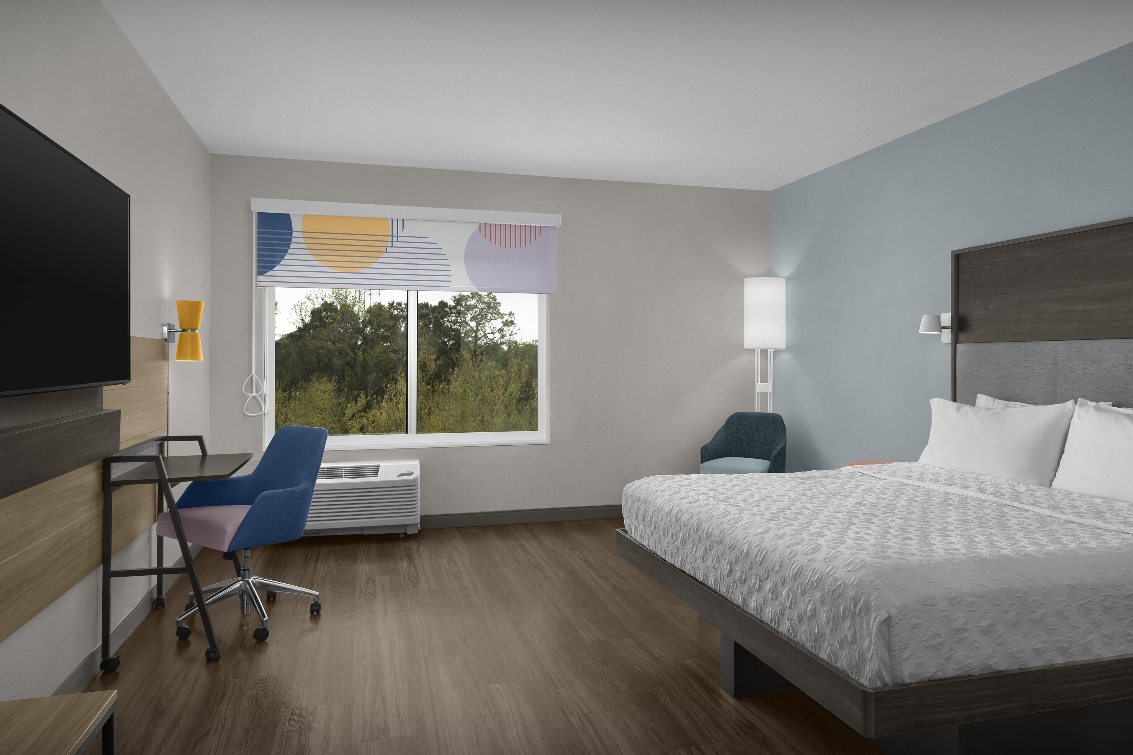 A hotel room with a bed, a desk, a chair, a TV, a window with blinds, and a lamp.